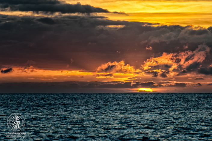 Golden sun dipping below dark ocean horizon at sunset.