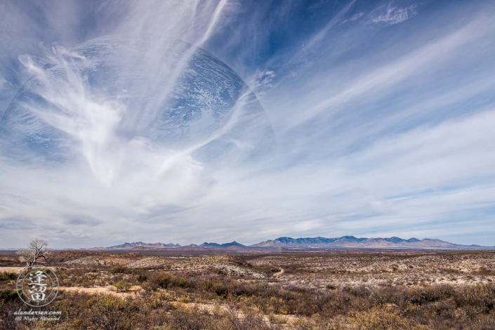 A digital composite of desert landscape with large planet in blue sky streaked by white wispy cirrus clouds.