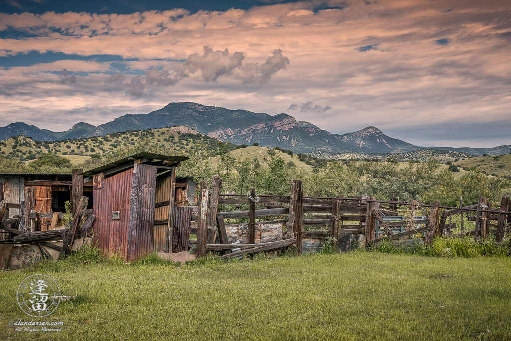 The outhouse and corrals at historic Brown Canyon Ranch during sunset on a warm August day with green grass in the foreground and Huachuca Peak in the Huachuca Mountains of Southeastern Arizona in the background.