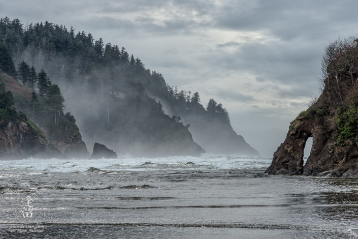 High tide beneath the misty hills on a cloudy day at Proposal Rock in Neskowin, Oregon.
