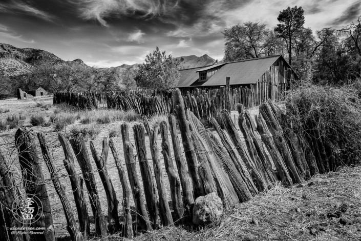 Stockaded barn surrounded by wire-woven log fence at historic Camp Rucker with adobe ruins in the distance.